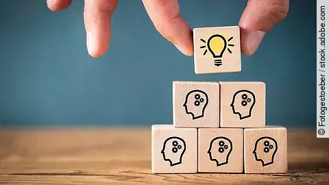Teamwork Fünf Holzwürfel sind übereinander gestapelt. Auf ihnen ist jeweils der Umriss eines Kopfes mit einem Fragezeichen zu sehen. Eine Hand legt ganz oben einen weiteren Würfel ab, auf dem eine Glühbirne abgebildet ist.