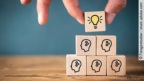 Fünf Holzwürfel sind übereinander gestapelt. Auf ihnen ist jeweils der Umriss eines Kopfes mit einem Fragezeichen zu sehen. Eine Hand legt ganz oben einen weiteren Würfel ab, auf dem eine Glühbirne abgebildet ist.