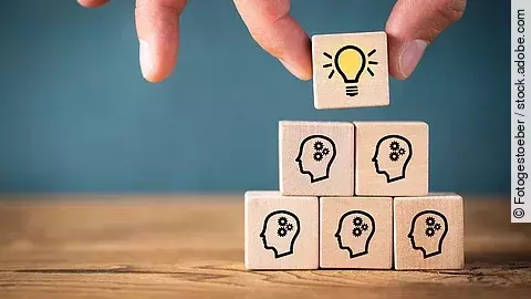 Teamwork Fünf Holzwürfel sind übereinander gestapelt. Auf ihnen ist jeweils der Umriss eines Kopfes mit einem Fragezeichen zu sehen. Eine Hand legt ganz oben einen weiteren Würfel ab, auf dem eine Glühbirne abgebildet ist.