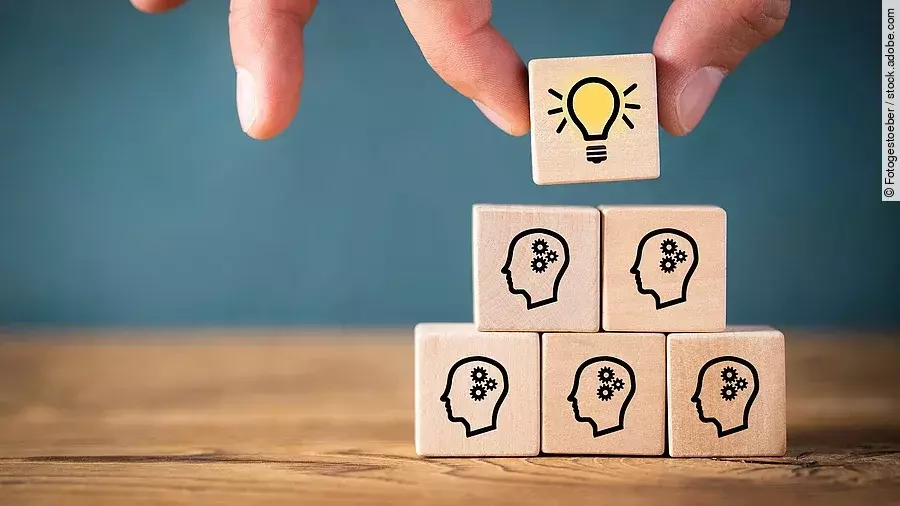 Fünf Holzwürfel sind übereinander gestapelt. Auf ihnen ist jeweils der Umriss eines Kopfes mit einem Fragezeichen zu sehen. Eine Hand legt ganz oben einen weiteren Würfel ab, auf dem eine Glühbirne abgebildet ist.