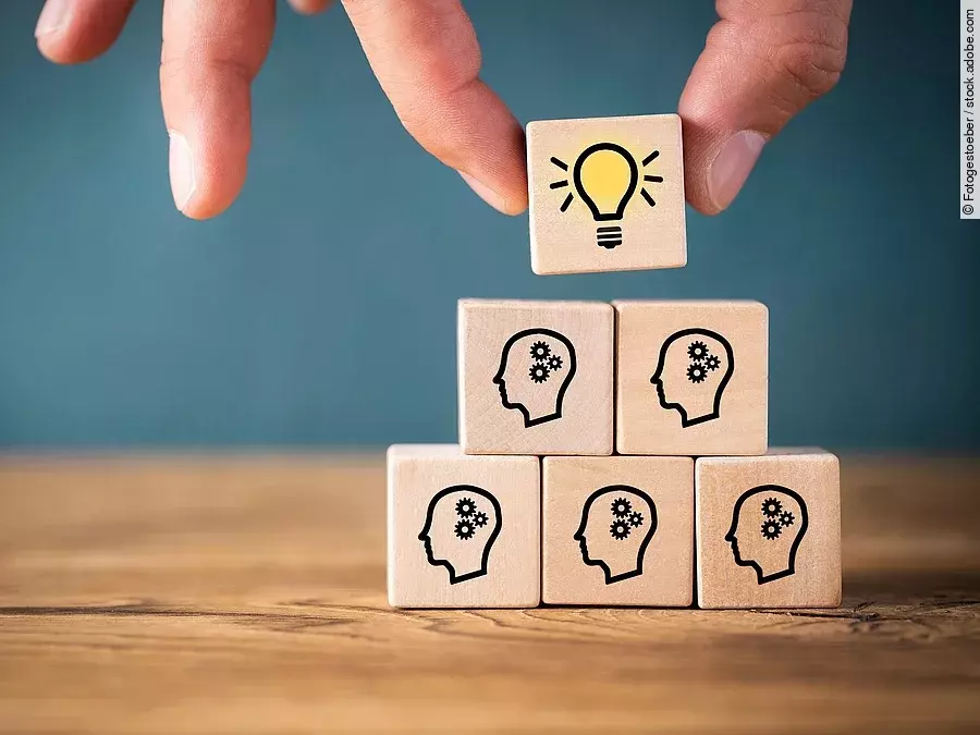 Teamwork Fünf Holzwürfel sind übereinander gestapelt. Auf ihnen ist jeweils der Umriss eines Kopfes mit einem Fragezeichen zu sehen. Eine Hand legt ganz oben einen weiteren Würfel ab, auf dem eine Glühbirne abgebildet ist.