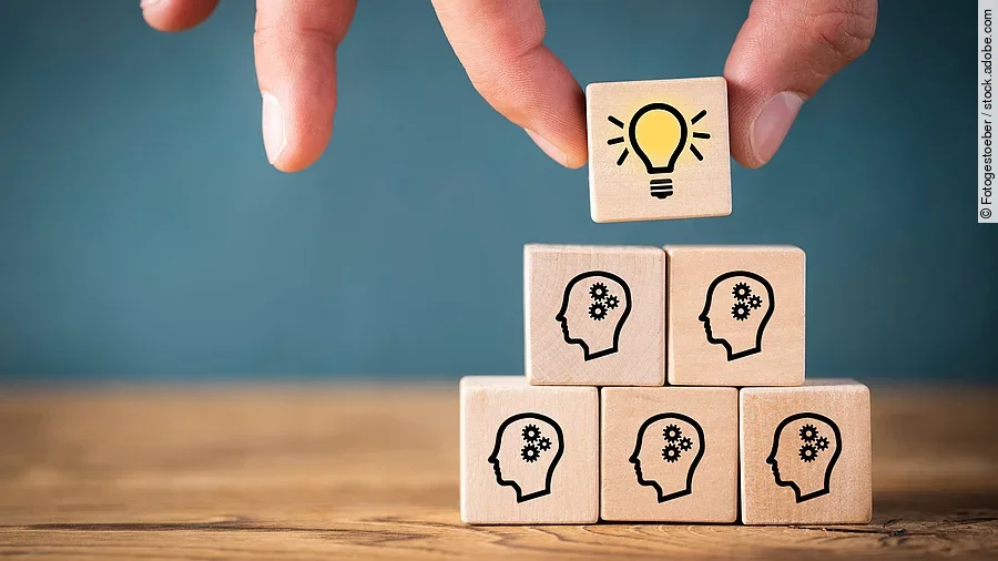 Teamwork Fünf Holzwürfel sind übereinander gestapelt. Auf ihnen ist jeweils der Umriss eines Kopfes mit einem Fragezeichen zu sehen. Eine Hand legt ganz oben einen weiteren Würfel ab, auf dem eine Glühbirne abgebildet ist.