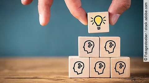 Teamwork Fünf Holzwürfel sind übereinander gestapelt. Auf ihnen ist jeweils der Umriss eines Kopfes mit einem Fragezeichen zu sehen. Eine Hand legt ganz oben einen weiteren Würfel ab, auf dem eine Glühbirne abgebildet ist.