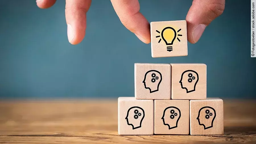 Fünf Holzwürfel sind übereinander gestapelt. Auf ihnen ist jeweils der Umriss eines Kopfes mit einem Fragezeichen zu sehen. Eine Hand legt ganz oben einen weiteren Würfel ab, auf dem eine Glühbirne abgebildet ist.