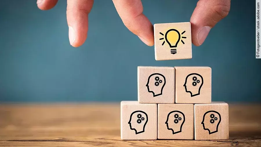 Fünf Holzwürfel sind übereinander gestapelt. Auf ihnen ist jeweils der Umriss eines Kopfes mit einem Fragezeichen zu sehen. Eine Hand legt ganz oben einen weiteren Würfel ab, auf dem eine Glühbirne abgebildet ist.