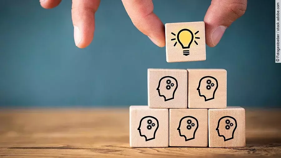 Teamwork Fünf Holzwürfel sind übereinander gestapelt. Auf ihnen ist jeweils der Umriss eines Kopfes mit einem Fragezeichen zu sehen. Eine Hand legt ganz oben einen weiteren Würfel ab, auf dem eine Glühbirne abgebildet ist.