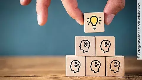 Teamwork Fünf Holzwürfel sind übereinander gestapelt. Auf ihnen ist jeweils der Umriss eines Kopfes mit einem Fragezeichen zu sehen. Eine Hand legt ganz oben einen weiteren Würfel ab, auf dem eine Glühbirne abgebildet ist.