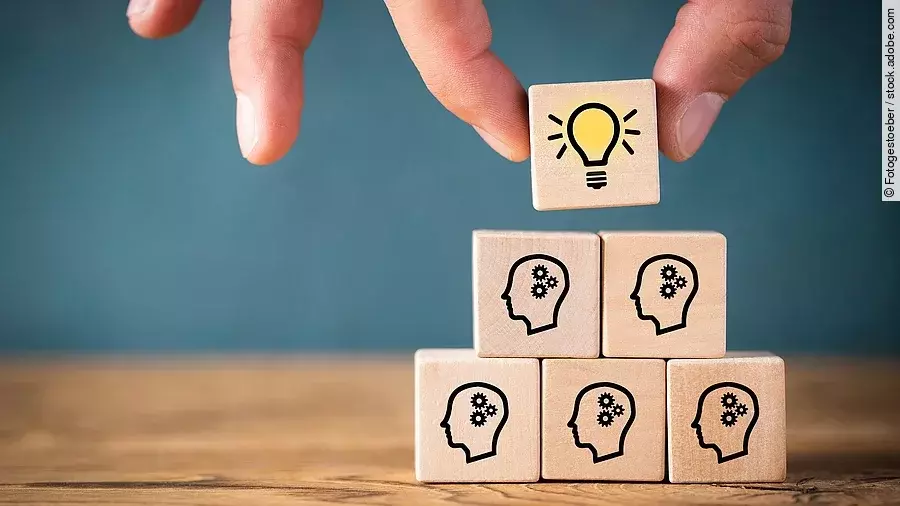 Fünf Holzwürfel sind übereinander gestapelt. Auf ihnen ist jeweils der Umriss eines Kopfes mit einem Fragezeichen zu sehen. Eine Hand legt ganz oben einen weiteren Würfel ab, auf dem eine Glühbirne abgebildet ist.