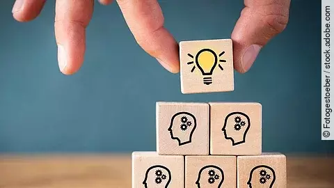 Fünf Holzwürfel sind übereinander gestapelt. Auf ihnen ist jeweils der Umriss eines Kopfes mit einem Fragezeichen zu sehen. Eine Hand legt ganz oben einen weiteren Würfel ab, auf dem eine Glühbirne abgebildet ist.