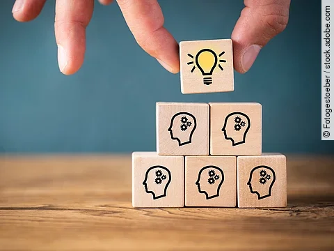 Fünf Holzwürfel sind übereinander gestapelt. Auf ihnen ist jeweils der Umriss eines Kopfes mit einem Fragezeichen zu sehen. Eine Hand legt ganz oben einen weiteren Würfel ab, auf dem eine Glühbirne abgebildet ist.