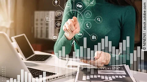 Frau in grünem Pullover sitzt vor einem Tisch mit Laptop und Dokumenten, die Grafiken zeigen. Mit einer Hand deutet sie auf in der Luft schwebende, imaginäre Symbole, die für Technologien und Daten stehen. Im Vordergrund sind zwei transparente Balkendiagramme abgebildet.