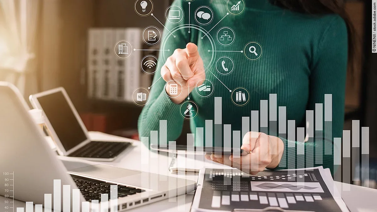 Frau in grünem Pullover sitzt vor einem Tisch mit Laptop und Dokumenten, die Grafiken zeigen. Mit einer Hand deutet sie auf in der Luft schwebende, imaginäre Symbole, die für Technologien und Daten stehen. Im Vordergrund sind zwei transparente Balkendiagramme abgebildet.
