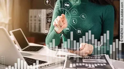 Frau in grünem Pullover sitzt vor einem Tisch mit Laptop und Dokumenten, die Grafiken zeigen. Mit einer Hand deutet sie auf in der Luft schwebende, imaginäre Symbole, die für Technologien und Daten stehen. Im Vordergrund sind zwei transparente Balkendiagramme abgebildet.