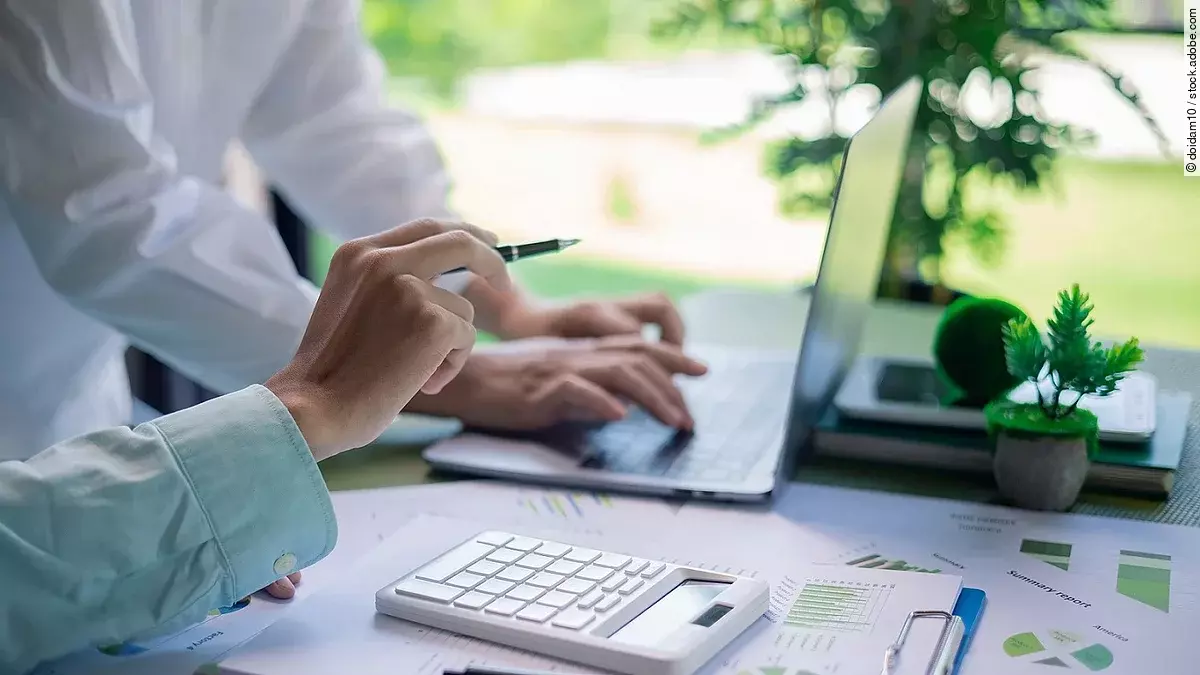 Zwei Personen in einer Besprechung. Vor ihnen ein aufgeklappter Laptop, Dokumente, Statistiken und ein Taschenrechner.