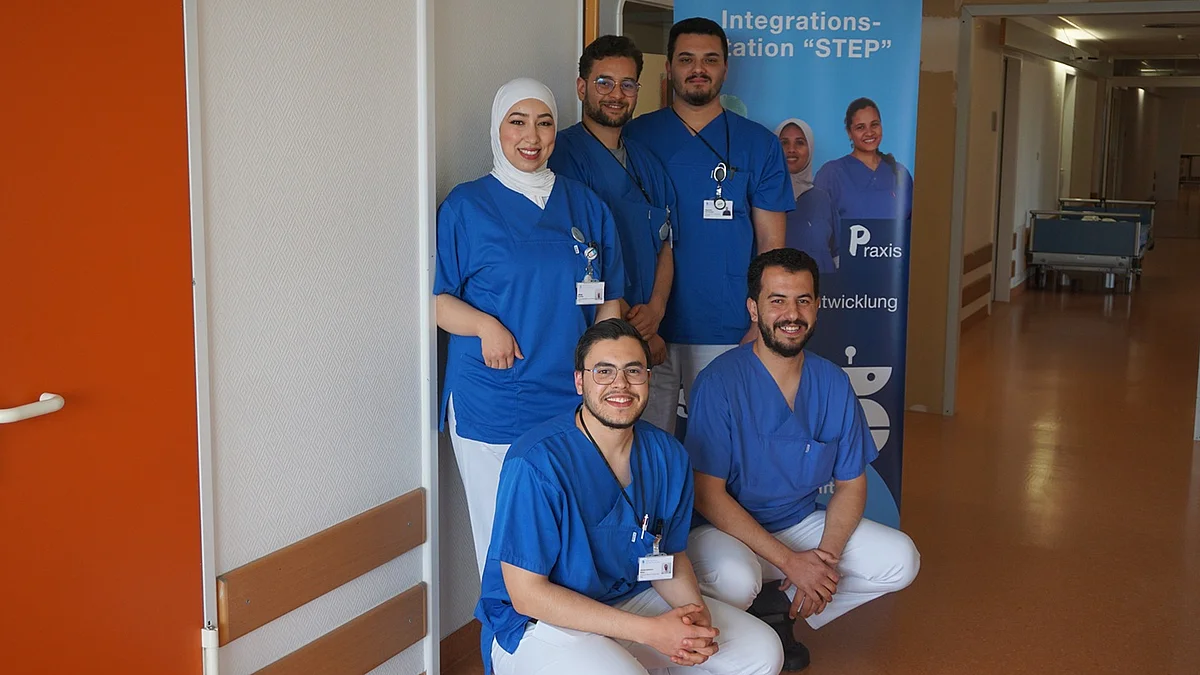 Step-Station im Sana Klinikum Hameln-Pyrmont Fünf Personen in blauen Kasacks und weißen Hosen, lächelnd, dahinter Aufsteller der Integrationsstation "Step"