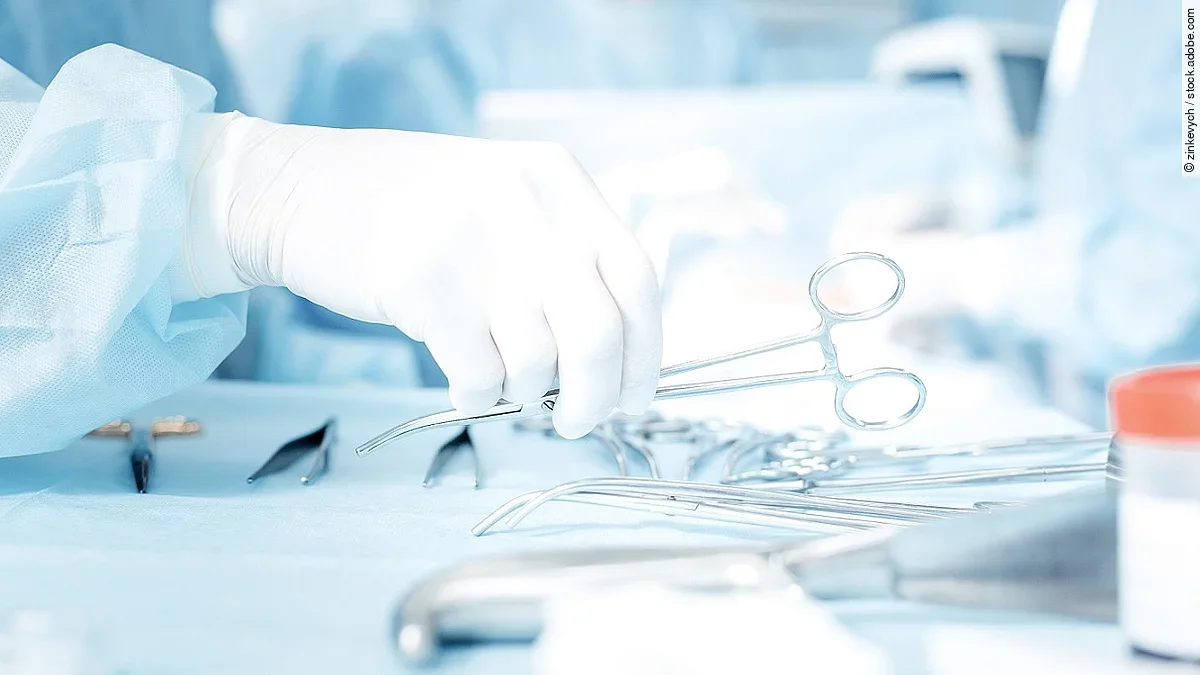 Chiurgie Hand greift nach OP-Schere, die auf einem Tisch mit weiterem OP-Besteck liegt.
