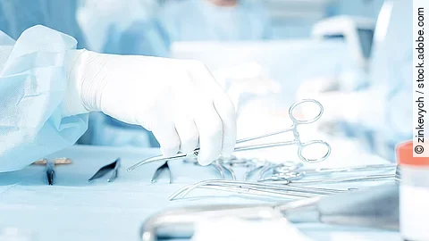 Hand greift nach OP-Schere, die auf einem Tisch mit weiterem OP-Besteck liegt.