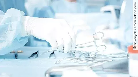 Hand greift nach OP-Schere, die auf einem Tisch mit weiterem OP-Besteck liegt.