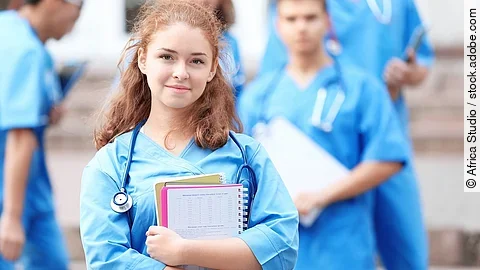 Medizinstudentin Eine Medizinstudentin blickt in die Kamera. Sie hat einen Block im Arm und ein Stethoskop um den Hals. Im Hintergrund sind weitere Studenten zu sehen.