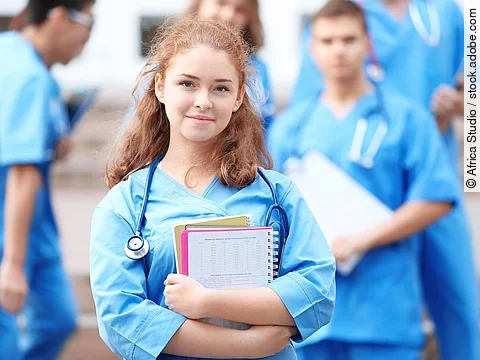 Eine Medizinstudentin blickt in die Kamera. Sie hat einen Block im Arm und ein Stethoskop um den Hals. Im Hintergrund sind weitere Studenten zu sehen.