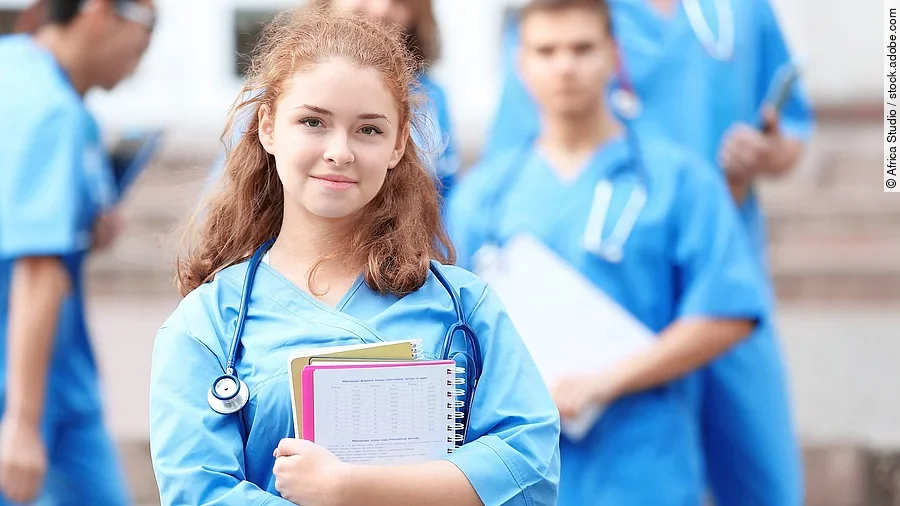 Eine Medizinstudentin blickt in die Kamera. Sie hat einen Block im Arm und ein Stethoskop um den Hals. Im Hintergrund sind weitere Studenten zu sehen.