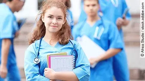 Eine Medizinstudentin blickt in die Kamera. Sie hat einen Block im Arm und ein Stethoskop um den Hals. Im Hintergrund sind weitere Studenten zu sehen.