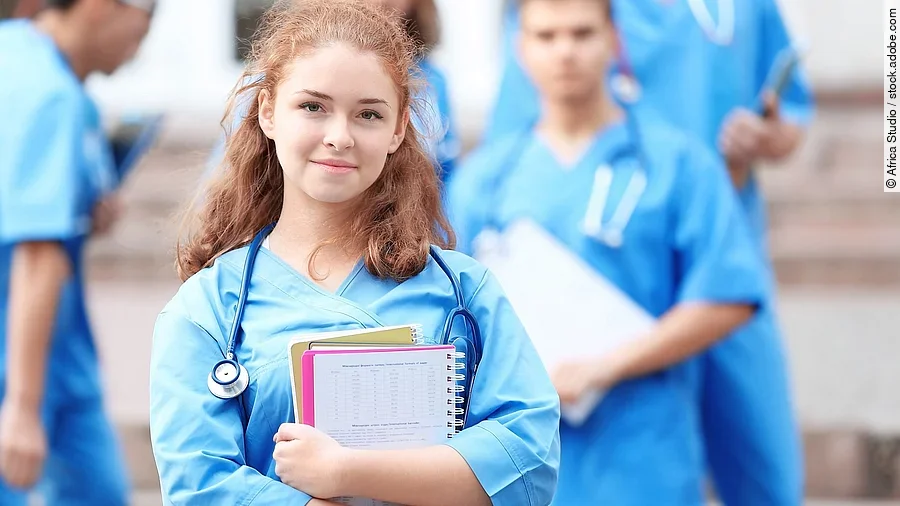 Medizinstudentin Eine Medizinstudentin blickt in die Kamera. Sie hat einen Block im Arm und ein Stethoskop um den Hals. Im Hintergrund sind weitere Studenten zu sehen.