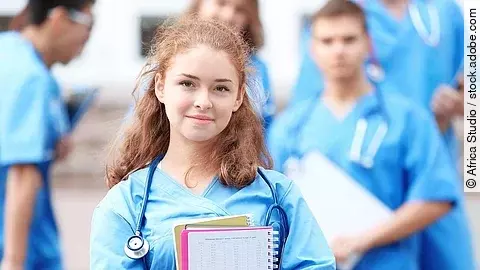 Eine Medizinstudentin blickt in die Kamera. Sie hat einen Block im Arm und ein Stethoskop um den Hals. Im Hintergrund sind weitere Studenten zu sehen.