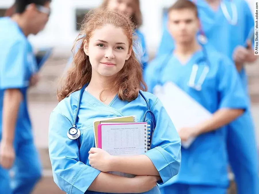 Eine Medizinstudentin blickt in die Kamera. Sie hat einen Block im Arm und ein Stethoskop um den Hals. Im Hintergrund sind weitere Studenten zu sehen.