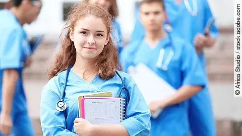 Eine Medizinstudentin blickt in die Kamera. Sie hat einen Block im Arm und ein Stethoskop um den Hals. Im Hintergrund sind weitere Studenten zu sehen.