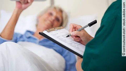 Eine Frau liegt im Pflegebett. Daneben steht eine Pflegekraft und dokumentiert.