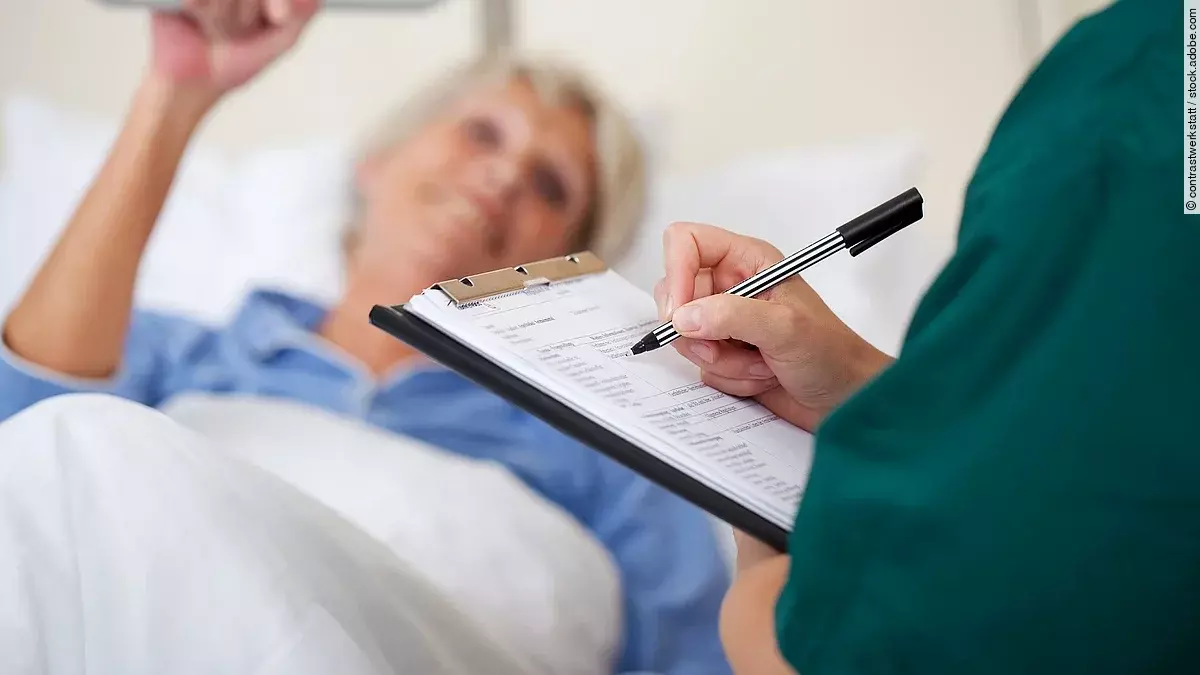 Entlassmanagement Eine Frau liegt im Pflegebett. Daneben steht eine Pflegekraft und dokumentiert.