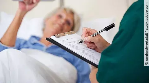 Eine Frau liegt im Pflegebett. Daneben steht eine Pflegekraft und dokumentiert.