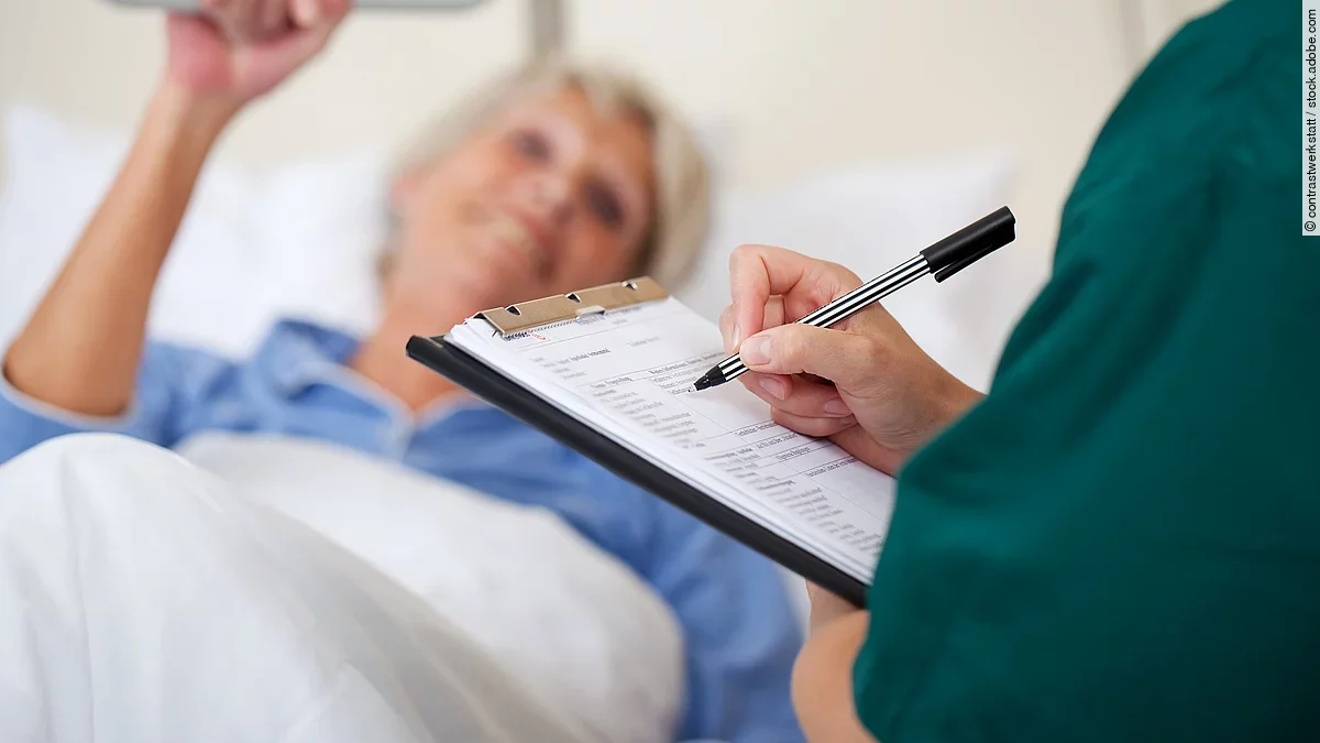 Eine Frau liegt im Pflegebett. Daneben steht eine Pflegekraft und dokumentiert.