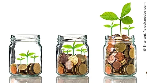 Investition Fünf Gläser stehen nebeneinander. Das linke ist leer, das rechte gänzlich mit Münzen gefüllt. Dazwischen steigt die Menge der Münzen. Aus den Münzen wächst eine Pflanze, die mit zunehmender Geldsumme immer größer wird.