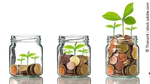 Fünf Gläser stehen nebeneinander. Das linke ist leer, das rechte gänzlich mit Münzen gefüllt. Dazwischen steigt die Menge der Münzen. Aus den Münzen wächst eine Pflanze, die mit zunehmender Geldsumme immer größer wird.