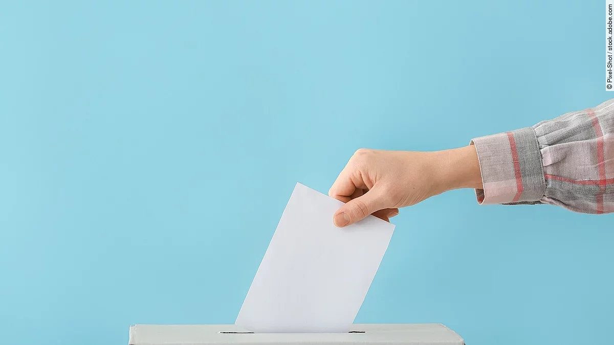 Wahl Ein Wahlzettel wird in eine graue Wahlurne geschmissen.